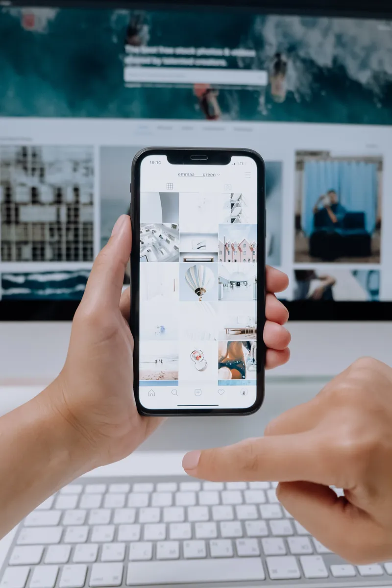 Manos sosteniendo un teléfono inteligente con la pantalla mostrando un perfil de Instagram con una variedad de publicaciones de diseño y arquitectura, frente a una computadora portátil con teclado y una persona desenfocada en el fondo